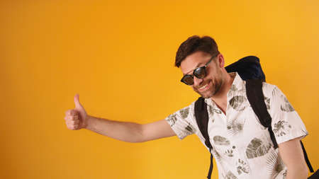 Young bearded man smiling with glasses and backpack holding thumb up, hitchhiking concept. High...