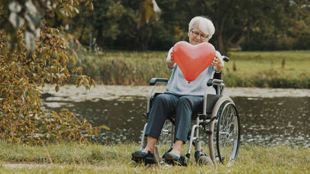 happy senior woman in the wheelchair near the river offering a balloon heart with a smile. Love and care concept. High quality photoの写真素材