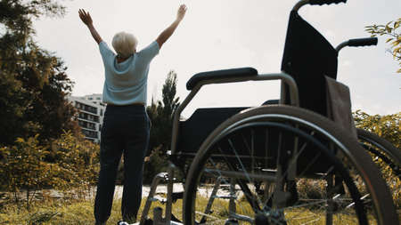 Recovery concept. Old woman with hands in the air in park in front of the wheelchair. High quality photoの写真素材