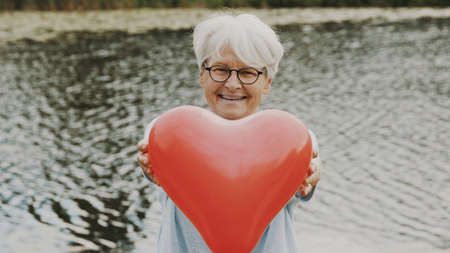Senior woman showing heart shaped balloon near the river. Love and care concept. High quality photoの写真素材