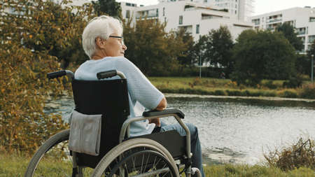 Senior disabled woman sitting in the wheelchair enjoying autumn breeze near the river. High quality photoの写真素材