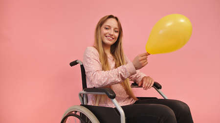 Young disabled woman in wheelchair holding yellow balloon, isolated on pink background. High quality photoの写真素材