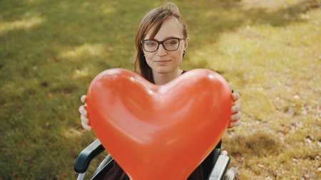 Beautiful young woman in the wheelchair holding heart shaped balloon in the park. High quality photo