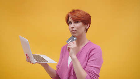 Confused redhead middle aged woman holding laptop and credit card. Online shopping concept. isolated...