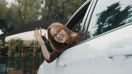 Happy face expression of young caucasian woman sitting on the driver seat and waving with car keys....
