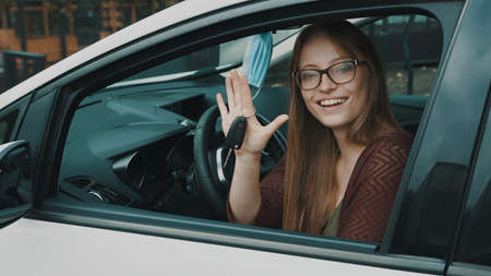 Happy face expression of young caucasian woman sitting on the driver seat and waving with car keys....