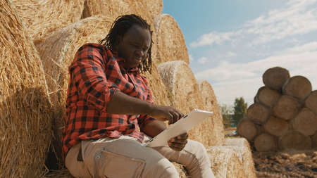 African black farmer sitting on the haystack and working on the laptop. High quality photo