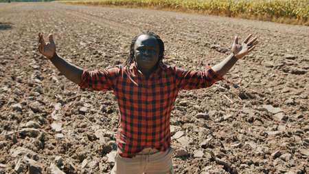 Happy african man, farmer looking around the plowed farmland with outstretched hands. High quality...