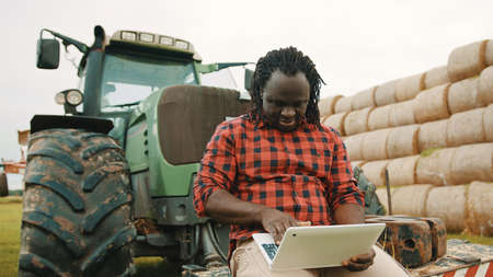 Young african farmer using tablet while sitting on green tractor. Haystack in the background. High...