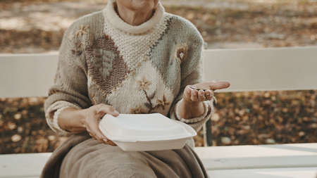 Homeless woman with charity meal and some coins in her hand sitting on the bench in autumn. High...