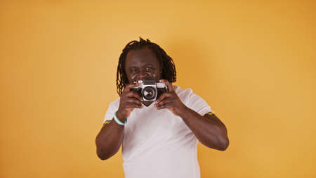 Portrait of an African man with braided hear and white shirt holding a vintage camera. Isolated on the orange background.の写真素材