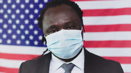 African American in Face Mask Looking at Camera - US Flag in Background - Close up. High quality photoの写真素材