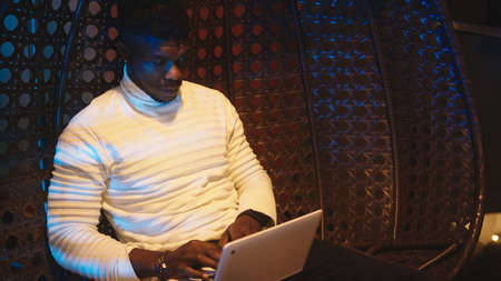 Young black man, freelancer sitting on the swing on the balcony and using laptop at night. High...