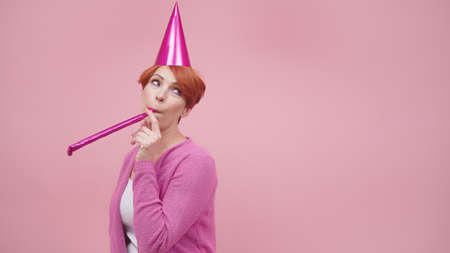 Bored red haired middle aged woman blowing party horn on the birthday party. Purple colors. High...