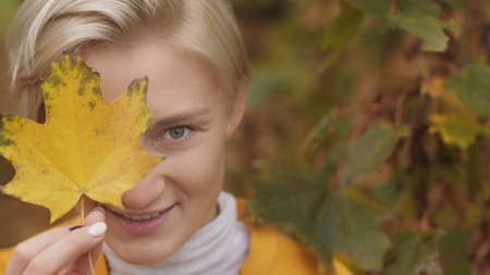 Portrait shot of young caucasian blonde woman with blue eyes hiding face with yellow autumn leaf....