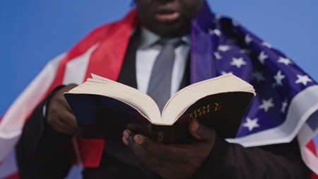 Close up, holy bible in the hands of black african american man in elegant suit with USA flag over...