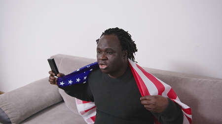African american black man with usa flag over the shoulders watching sport game on tv on his couch....