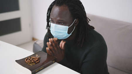 Black African Descent Man With Face Mask Praying on Holy Bible Book and Rosary. Seeking Hope in...