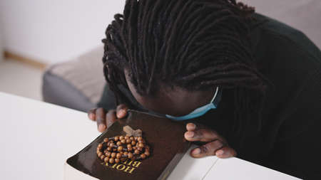 Black African Descent Man With Face Mask Praying on Holy Bible Book and Rosary. Seeking Hope in...