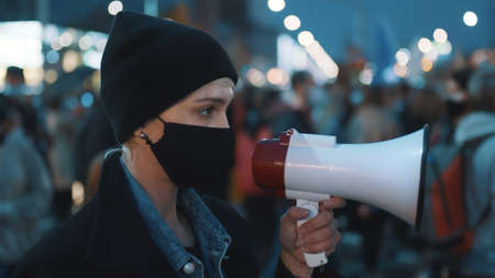 young short haired woman with medical mask on face shout out slogans to loudspeaker at street...