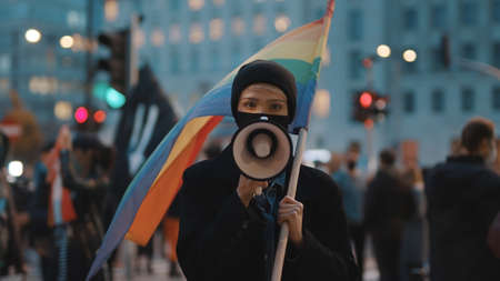 Young woman with face mask speaking into the megaphone while holding rainbow flag in crowd. High quality photoの写真素材