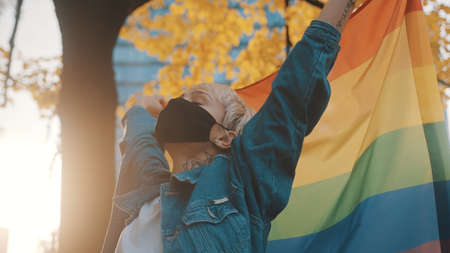 Proud young blond woman with face mask waving rainbow flag. Close up. High quality photoの写真素材