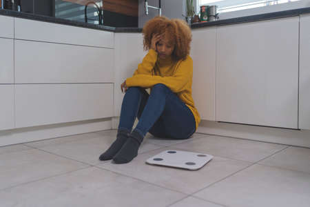 Devastated young african american black woman sitting on the flor with weight scale in front. Weight...