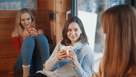 Winter holidays. Girls having fun in the cozy wooden hut on the mountain. Drinking hot beverage near...