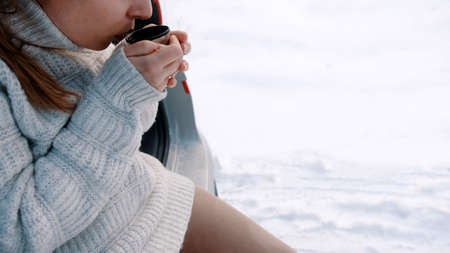 Close up, young woman leaning on the car and drinking hot beverage on the winter snowy day. High...