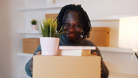 Excited young african american man moving in to the new apartment. Holding cardboard boxes with...