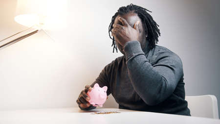 Empty piggy bank. Sad african american man putting one coin inside. Small savings. High quality photoの写真素材