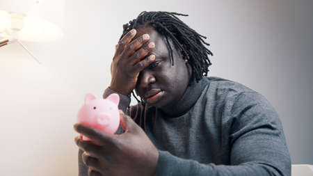 Bankruptcy concept. Sad african american black man looking at empty piggy bank and shaking his head....
