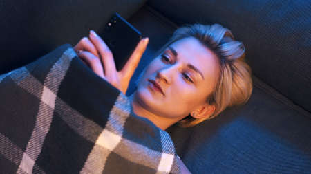 Portrait of young woman browsing social media while relaxing on the sofa covered with blanket. High...