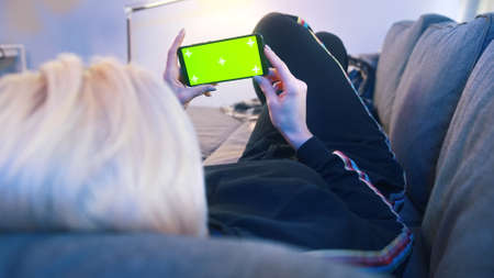 Young woman relaxing on the couch and holding smartphone with green screen. High quality photo