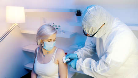 Nurse in full protective uniform giving vaccie to young blonde woman. High quality photo