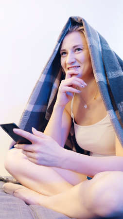 Young caucasian woman covered with blanket using smartphone . High quality photo