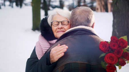 Valentines day. Happy elderly man giving bouquet of red roses to his date in winter. Hugging un the...