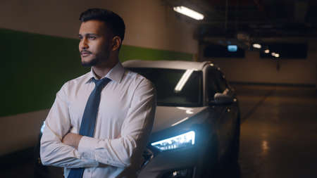 Waist up of adult businessman standing indoors in front of his car with arms crossed and looking...