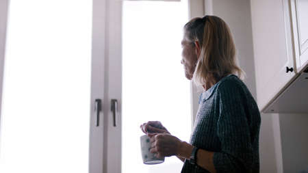 Lonely elderly woman drinking coffee standing at her kitchen. High quality photo