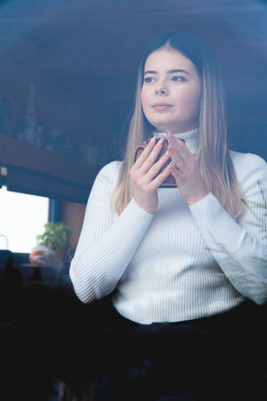 Girl holding coffee mug looking through a window. Wearing a white high-neck shirt and black pants....
