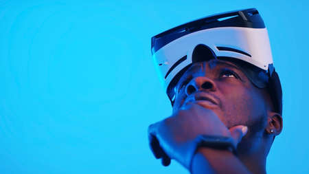 Pensive African-American guy with virtual reality glasses on his forehead looking around. High...