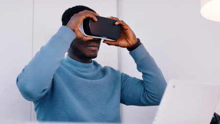 Black man wearing a virtual reality glasses sitting at desk with his laptop. High quality photoの写真素材