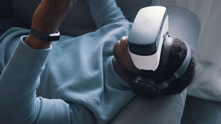 African-American man wearing virtual reality glasses and headset lying on the sofa. Man working...