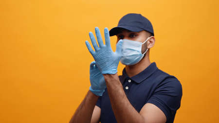 Delivery man putting on the gloves. Wearing a mask and a blue cap taking precautions. Concept of Covid 19 and Pandemic. Yellow background studio shot.の写真素材
