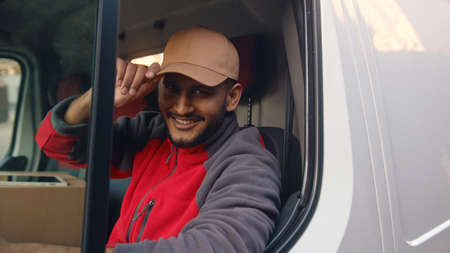 Delivery guy wearing red uniform sitting in the white van and tipping his cap. Happy Indian guy sitting in the front seat and smiling at the camera. Parcel delivery concept.の写真素材