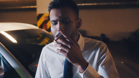 Young man in the parking lot smoking. Dressed in white shirt and tie. White car behind him. Stressed out man with a cigarette in his hand. Concept of Work load and depression.の写真素材