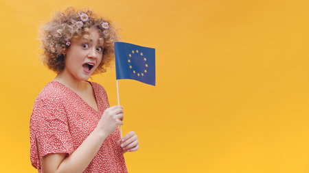 Beautiful young girl holding a small European Union Flag in her hand. Astonished girl wearing casual pink top. Studio shot isolated on yellow background. Symbol of Europe, EU association.の写真素材