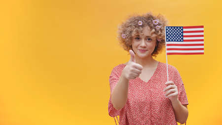 Attractive young girl holding a small USA flag. Smiling and making thumbs up sign at the camera....