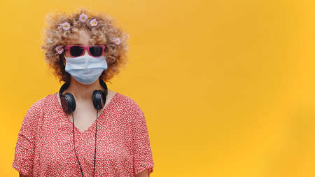 Beautiful charming girl wearing sunglasses and a disposable face mask. Headphones around her neck. Dressed in pink casual top. Isolated over bright yellow background studio.の写真素材