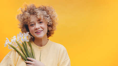 Attractive girl holding a bunch of white spring Flower in her hands. Girl with curly hair smiling and dressed in a baggy yellow sweater. Isolated over bright yellow background studio. Springtime.の写真素材